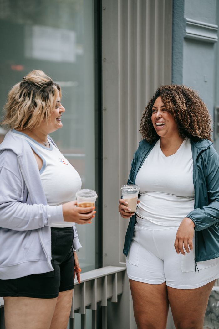 Two women in sportswear laughing and holding iced drinks outside on a street.